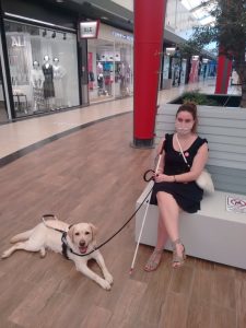 Η Χριστίνα Σαρρή με ροζ μάσκα και μαύρο φόρεμα, καθισμένη σε ένα παγκάκι. Μπροστά της βρίσκεται ο σκύλος οδηγός της