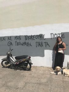 Η Χριστίνα με τη Luna, το σκύλο οδηγό της, μπροστά από έναν τοίχο στο Βόλο που γράφει: «Κι ας μη νικήσουμε ποτέ, θα πολεμάμε πάντα»