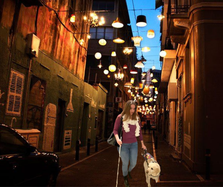 Βραδινή φωτογραφία με τη Χριστίνα Σαρρή και το σκύλο οδηγό της να περπατούν σε ένα στενό. Η Χριστίνα κρατάει ένα μικρό, λευκό μπαστούνι στο δεξί της χέρι κι έχει μια μαύρη τσάντα περασμένη στον ώμο.