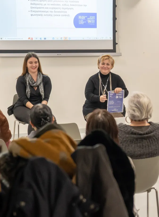 Αίθουσα συνεδριάσεων. Κόσμος καθισμένος σε καρέκλες και στο βάθος μιλάνε η Αρετή Βάσσου και η Χριστίνα Σαρρή.
