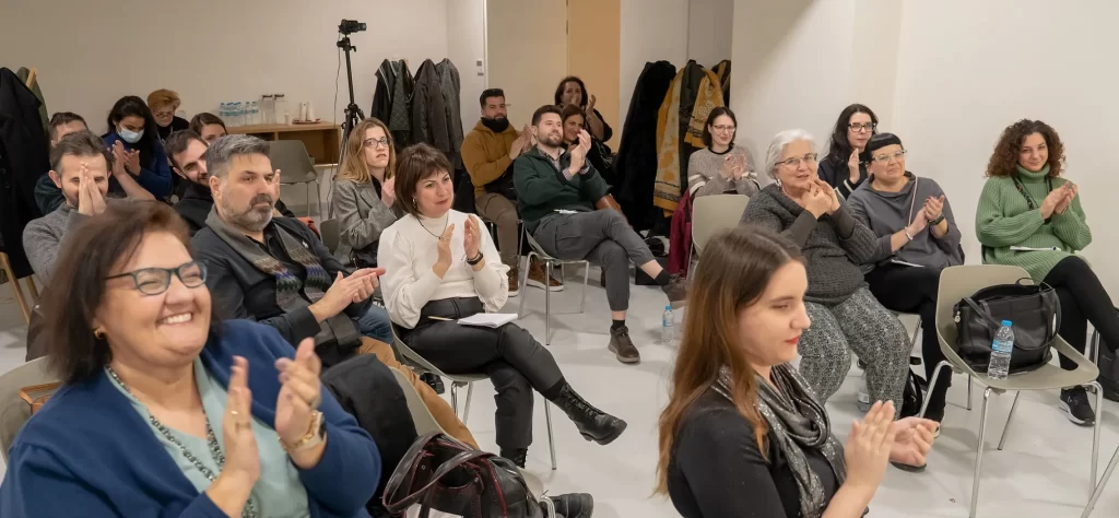 Γενική φωτογραφία του κόσμου που παρακουλουθεί το meetup, μέσα στην αίθουσα συνεδριάσεων. Φαίνονται πολλά και διαφορετικά πρόσωπα. Ο κόσμος χειροκροτεί.
