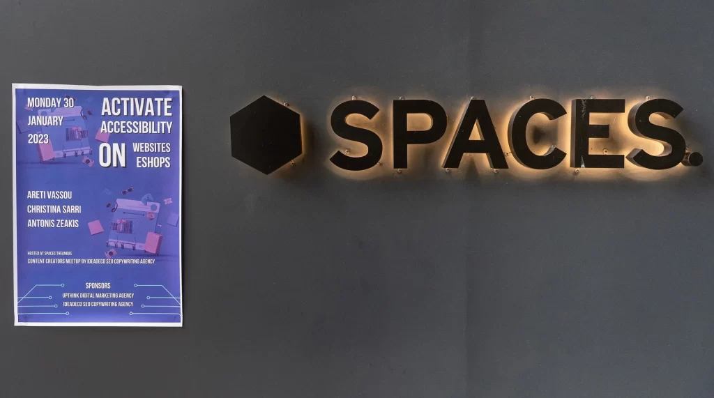 Αφίσα του meetup πάνω σε γκρι τοίχο, δίπλα στην επωνυμία του χώρου SPACES. Στο κέντρο είναι η κεντρική διπλή πόρτα εισόδου. Δεξιά φαίνεται κομμάτι από τα παράθυρα του χώρου.