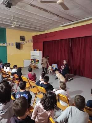 Τα παιδιά καθισμένα σε καρέκλες, παρακολουθούν τη Χριστίνα Σαρρή που έχει σηκωθεί όρθια και σκύβει να πιάσει τη Λούνα από το ειδικό σαμάρι. 
Πιο δίπλα είναι καθισμένη η κυρία Ζωή Γερουλάνου μαζί με την Ίρμα.