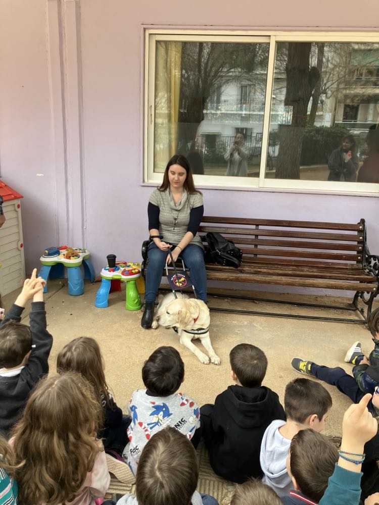 Η Χριστίνα Σαρρή  μιλάει μπροστά σε παιδιά για τους σκύλους οδηγούς. Κοντά της κάθεται η λούνα φορώντας το γιλέκο εργασίας της.
