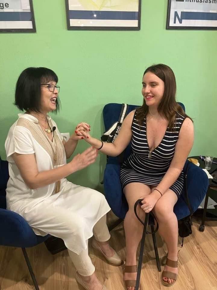 Η Χριστίνα Σαρρή κι η Γώνη Μούκα καθισμένες δίπλα δίπλα στην αίθουσα του σεμιναρίου. Έχουν πλεγμένα τα χέρια τους σφιχτά σε μία κοινή γροθιά και κοιτιούνται χαμογελαστές.