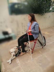 Η Χριστίνα Σαρρή είναι καθιστή και κρατάει μπαστούνι. Η Λούνα κάθεται μπροστά από την Χριστίνα Σαρρή λοξά και έχει σταυρωμένα τα πόδια της.