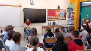 Σε σχολική αίθουσα με μαυροπίνακα φαίνεται η Χριστίνα με τη Λούνα στο βάθος και μπροστά της βρίσκονται τα παιδιά που παρακολουθούν την ομιλία της με την πλάτη γυρισμένη στο φακό.