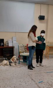  Η Χριστίνα Σαρρή στέκεται όρθια και με τη βοήθεια ενός μαθητή εξηγεί τις τεχνικές συνοδείας ενός τυφλού ατόμου. Ο μαθητής έχει λυγισμένο το δεξί του χέρι κι η Χριστίνα τον κρατά από το μπράτσο. Η Λούνα περιμένει ήρεμη πίσω, κοντά στην κενή καρέκλα.