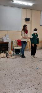 Η Χριστίνα Σαρρή στέκεται όρθια και με τη βοήθεια ενός μαθητή εξηγεί τις τεχνικές συνοδείας ενός τυφλού ατόμου. Κρατά το μαθητή από το μπράτσο. Η Λούνα περιμένει ήρεμη πίσω, κοντά στην κενή καρέκλα.