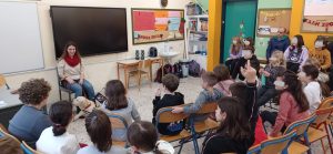  Σε σχολική αίθουσα με μαυροπίνακα φαίνεται η Χριστίνα με τη Λούνα στο βάθος και μπροστά της βρίσκονται τα παιδιά που παρακολουθούν την ομιλία της με την πλάτη γυρισμένη στο φακό. 