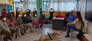 Μέσα σε σχολική αίθουσα η Χριστίνα Σαρρή δίνει ομιλία και κάποια παιδιά σηκώνουν το χέρι τους για να κάνουν ερωτήσεις. Η Λούνα, φορώντας το γιλέκο της, έχει γυρισμένη την πλάτη προς τα παιδιά και κοιτάζει τη Χριστίνα. Στην έδρα δίπλα τους βρίσκεται μια δασκάλα και πίσω απ’ τη Χριστίνα υπάρχει ένας μαυροπίνακας. 