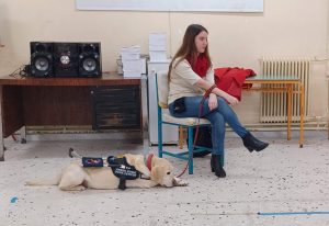 Η Χριστίνα Σαρρή καθισμένη σε καρέκλα με τη Λούνα στα δεξιά της. Η Λούνα είναι ξαπλωμένη και φοράει το ειδικό γιλέκο που δηλώνει ότι είναι σκύλος οδηγός.