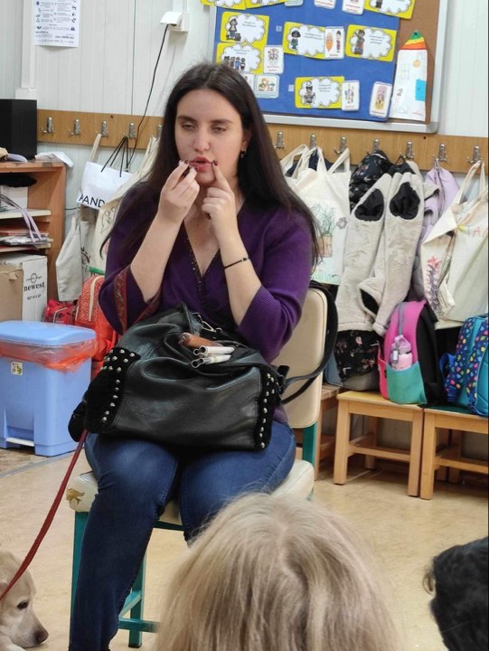 Η Χριστίνα Σαρρή με τη Λούνα ξαπλωμένη κοντά της δείχνει σε παιδιά νηπιαγωγείου πώς φοράει το κραγιόν της. Επάνω στα πόδια της βρίσκεται η τσάντα της με το λευκό μπαστούνι διπλωμένο από πάνω.