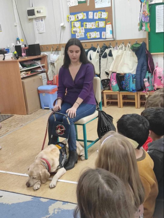 Η Χριστίνα Σαρρή καθισμένη δίνει ομιλία σε σχολείο. Η Λούνα είναι ξαπλωμένη στα πόδια της φορώντας το γιλέκο εργασίας της. Αριστερά φαίνονται μαθητές και μαθήτριες και πίσω απ’ τη Χριστίνα υπάρχουν σχολικές τσάντες.