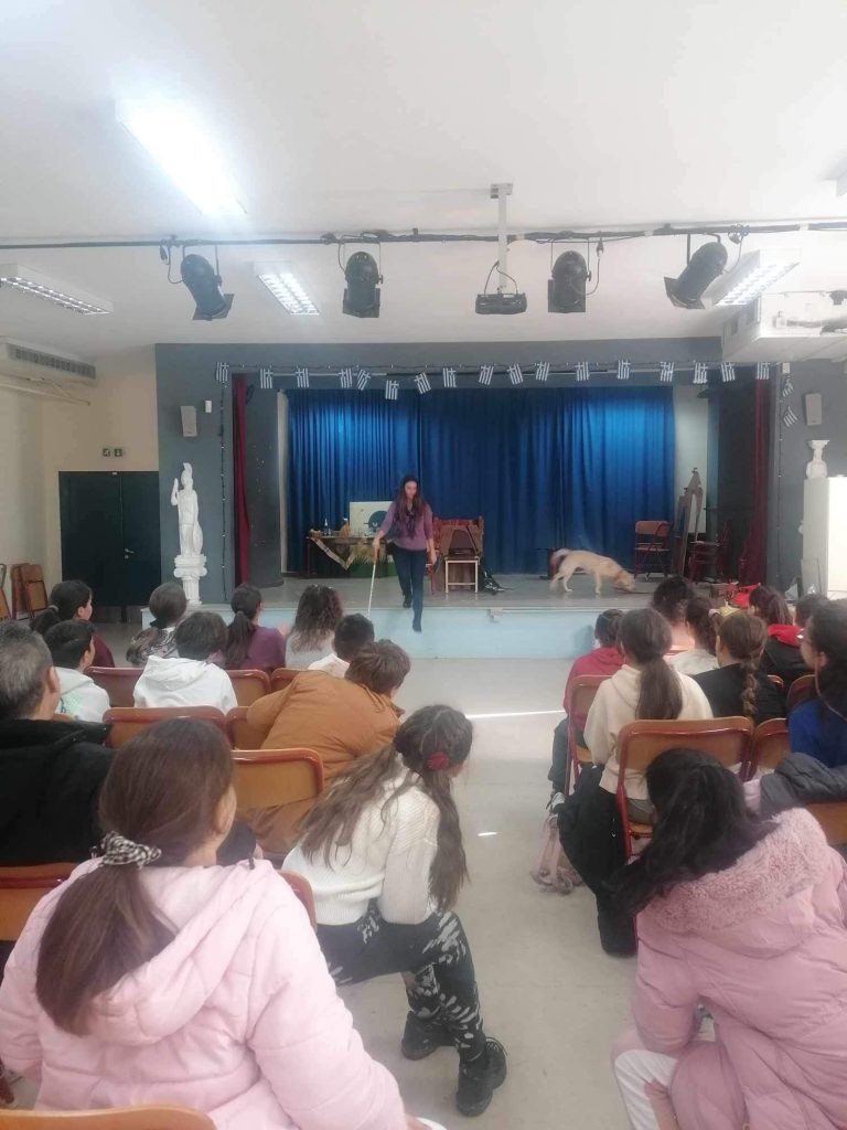 Η Χριστίνα Σαρρή επάνω σε εξέδρα αίθουσας εκδηλώσεων δημοτικού σχολείου. Πολλά παιδιά την παρακολουθούν με προσοχή να τους μιλά για το λευκό μπαστούνι, ενώ η Λούνα εξερευνά το χώρο μυρίζοντας το πάτωμα. Πίσω υπάρχουν μπλε κουρτίνες και από πάνω κρέμονται ελληνικές σημαίες.