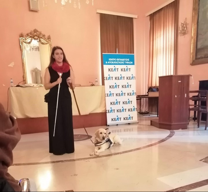 Η Χριστίνα Σαρρή όρθια δίνει ομιλία σε ενήλικο κοινό. Κρατάει ένα λευκό μπαστούνι σύμβολο και αριστερά της είναι ξαπλωμένη η Λούνα, ο σκύλος οδηγός της. Βρίσκονται σε αίθουσα πνευματικού κέντρου.